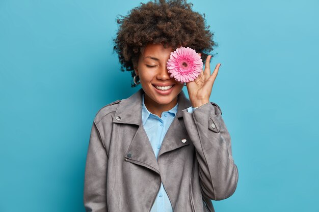 La imagen interior de una floristería profesional cubre el ojo con gerbera daisy, hace una composición creativa de flores, prepara el ramo para la venta, sonríe ampliamente, se coloca