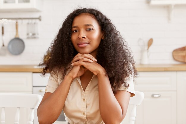 Imagen interior de una hermosa joven negra de piel oscura con peinado afro y piel bronceada bronceada cocinando en la cocina, sentada a la mesa, manteniendo las manos juntas debajo de la barbilla, mirando
