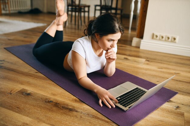 Imagen interior de hermosa mujer joven con cuerpo musculoso en forma acostada boca abajo sobre una estera de yoga navegando por internet a través de una computadora portátil después del entrenamiento en casa.