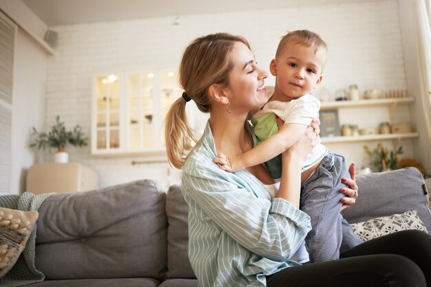 Imagen interior de linda hembra joven con cola de caballo sosteniendo apretado a su encantador bebé, sentada en el sofá con él. Bonita madre e hijo que se unen en la sala de estar, mamá mirando al niño con amor y ternura