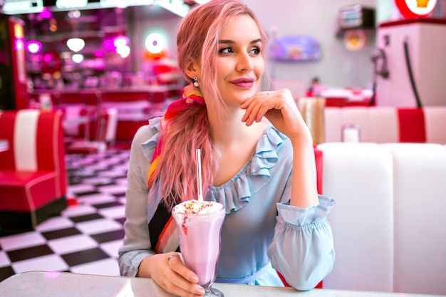 Imagen interior de una mujer elegante bastante joven que disfruta de su sabroso batido de fresa dulce en un restaurante americano retro vintage, diseño de neón, lindo vestido pastel, pelos rosados y accesorios