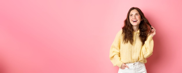 Foto gratuita imagen de una joven de 20 años riéndose y jugando con su cabello mirando a un lado la promoción de la esquina superior izquierda de pie feliz contra un fondo rosa