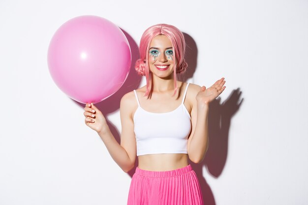 Imagen de una joven alegre con peluca rosa y maquillaje brillante, sosteniendo un globo y agitando la mano para saludar a alguien en la fiesta, celebrando la fiesta, blanco.