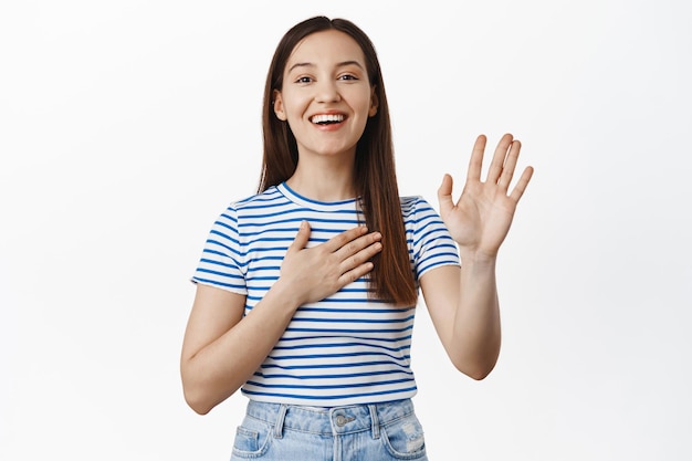 Imagen de una joven alegre que se presenta, se ofrece como voluntaria, levanta la mano y sostiene la palma en el corazón, promete, sonríe y se ve feliz, quiere participar en algo, autopromocionarse