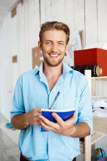 Imagen de joven apuesto hombre de negocios sonriente confiado escribiendo en el cuaderno. Interior de la oficina moderna blanca