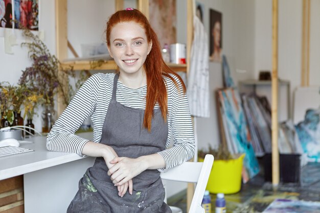 Imagen de una joven artesana talentosa con cara bonita y linda sonrisa con un delantal sucio con pinturas descansando después de terminar el trabajo, sentada en una silla en el interior del taller creativo moderno