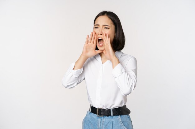 Imagen de una joven asiática llamando a alguien gritando fuerte y buscando de pie contra un fondo blanco