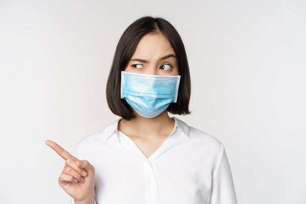 Imagen de una joven asiática con mascarilla médica apuntando a la izquierda y mirando con una expresión sospechosa y confusa de pie sobre fondo blanco