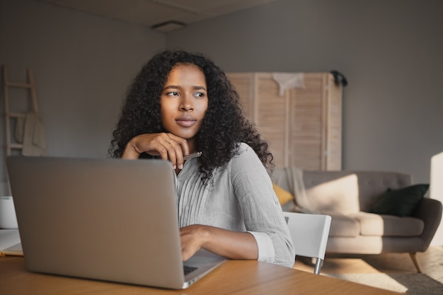 Imagen de la joven y atractiva mujer de piel oscura independiente de moda en camisa sentado en el lugar de trabajo en casa y usando conexión inalámbrica a Internet de alta velocidad en la computadora portátil, con mirada pensativa