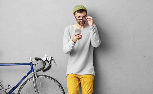 Imagen de un joven barbudo con un sombrero de moda y ropa con anteojos mientras lee un mensaje de texto extraño de un número desconocido, mira la pantalla, tiene una mirada sospechosa o desconfiada
