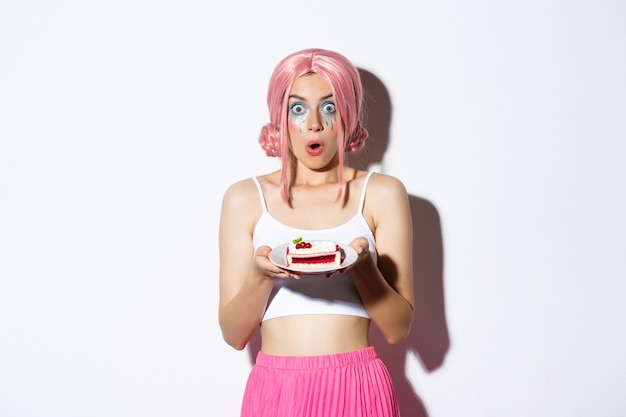 Imagen de la joven cumpleañera sorprendida con peluca rosa, sosteniendo el pastel en un plato y mirando asombrado a la cámara, de pie sobre fondo blanco.