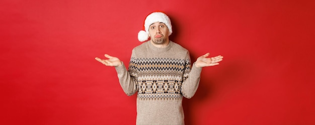 Imagen de un joven despistado con suéter y gorro de Papá Noel, encogiéndose de hombros y sin darse cuenta, no sabe qué comprar para las vacaciones de año nuevo, de pie sobre un fondo rojo