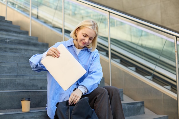 Foto gratuita imagen de una joven elegante guardando su computadora portátil en una mochila sentada en las escaleras cerca del campus