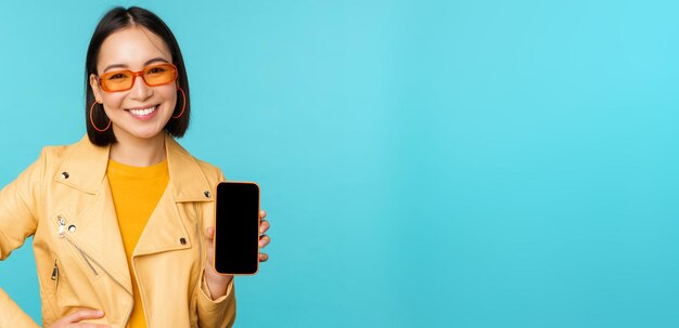 Imagen de una joven y elegante modelo asiática con gafas de sol de moda que muestra una aplicación de pantalla de teléfono móvil en la interfaz de un smartphone de pie sobre un fondo azul