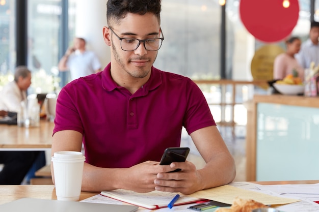 Imagen de un joven elegante que sostiene un teléfono móvil, recibe una notificación de ingresos, verifica el buzón de correo electrónico, está conectado a Internet de alta velocidad, usa anteojos ópticos, desarrolla un proyecto de investigación y asigna una casa