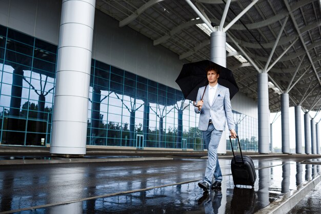 Imagen del joven empresario pelirrojo con paraguas negro y maleta caminando bajo la lluvia en la estación