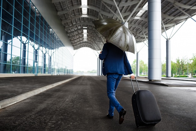 Imagen del joven empresario sosteniendo la maleta y el paraguas en el aeropuerto lluvioso