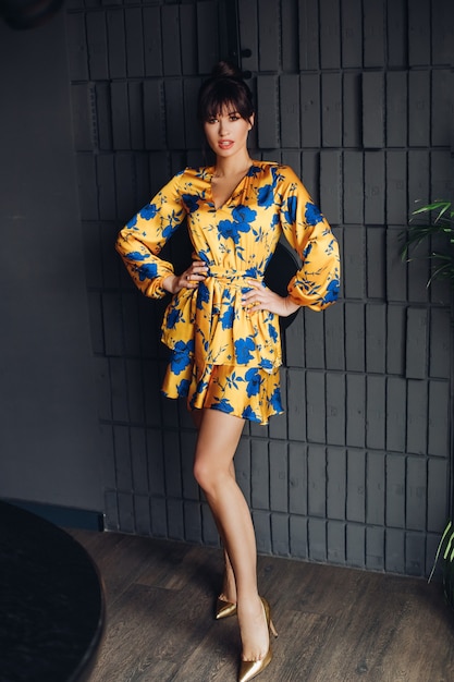 Imagen de joven encantadora mujer caucásica con cabello oscuro en vestido amarillo y azul, zapatos dorados muestra diferentes poses para la cámara