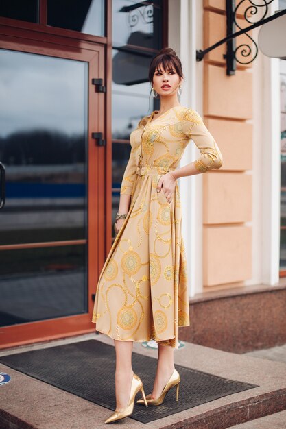 Imagen de joven encantadora mujer caucásica con cabello oscuro en vestido dorado y zapatos dorados muestra diferentes stands cerca del hermoso edificio