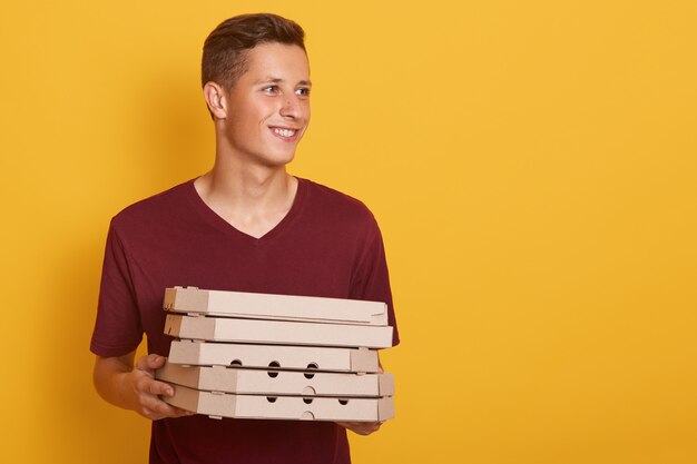 Imagen de un joven enérgico positivo con camiseta roja informal, sosteniendo cajas de pizza de cartón en ambas manos, mirando a un lado, sonriendo sinceramente, de buen humor. Copyspace para publicidad.