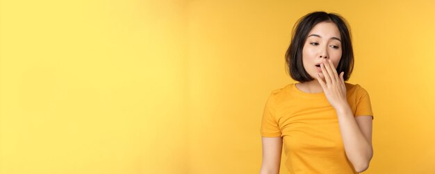 Imagen de una joven estudiante asiática bostezando sintiéndose cansada de pie con sueño contra un fondo amarillo