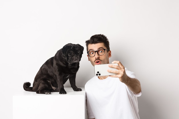 Imagen de joven guapo tomando selfie con lindo perro negro en smartphone, posando con pug sobre fondo blanco.