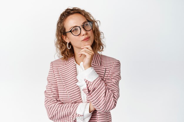 Imagen de la joven jefa con gafas y traje pensando, toma una decisión, toca la barbilla y se ve pensativa en blanco.