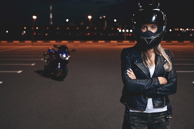 Imagen de una joven jinete segura de sí misma con casco de seguridad de pie en el estacionamiento con los brazos cruzados y mirando, yendo a andar en motocicleta por la ciudad de noche