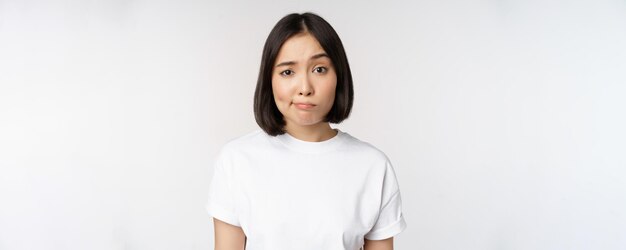 Imagen de una joven morena asiática con camiseta blanca sonríe y mira con duda dudando de pie contra el fondo blanco Copiar espacio