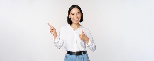 Imagen de una joven mujer de negocios asiática sonriendo mientras señala con el dedo a la izquierda y muestra el pulgar hacia arriba recomendando elogios del producto de pie sobre fondo blanco