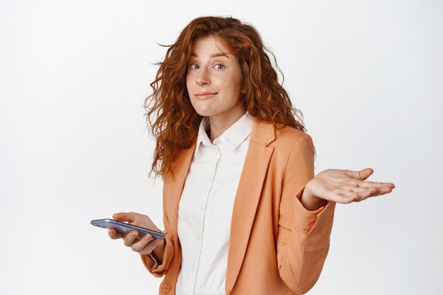 Imagen de una joven mujer de negocios con traje sosteniendo un teléfono móvil y encogiéndose de hombros, despistada, desconcertada y sonriendo para disculparse con el fondo blanco