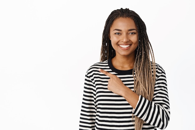 Imagen de una joven negra sonriente señalando con el dedo a la izquierda mostrando una venta promocional de espacio de copia de pie sobre un fondo blanco