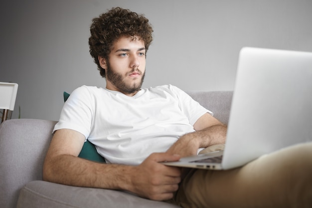 Imagen de un joven periodista caucásico confiado en ropa casual que tiene una mirada enfocada mientras trabaja de forma remota en una computadora portátil, haciendo una investigación, acostado cómodamente en el sofá con un dispositivo en su regazo