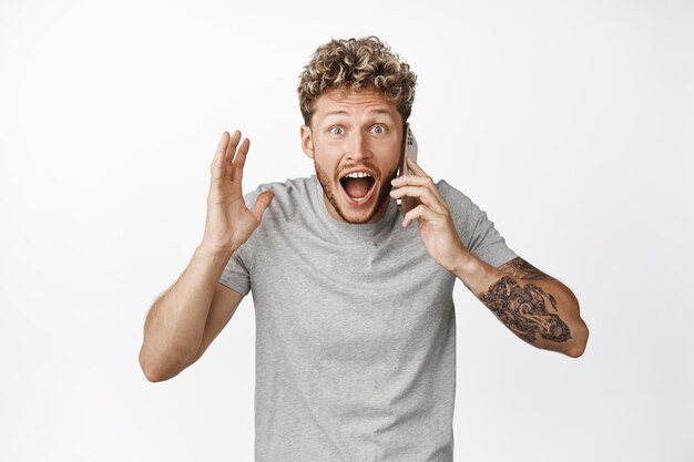 La imagen de un joven que habla por teléfono recibe una llamada telefónica y mira con incredulidad gritando a la cámara con una expresión de cara emocionada de fondo blanco