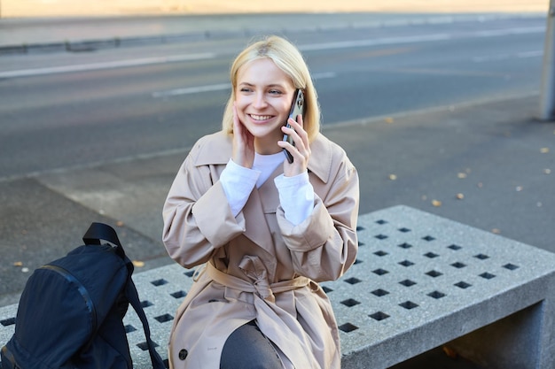Foto gratuita la imagen de una joven rubia sentada en un banco de la calle con una mochila hablando por teléfono móvil responde a una