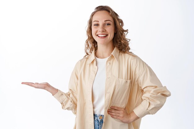 La imagen de una joven rubia sostiene un artículo en la mano y muestra su logotipo en la palma de la mano contra el espacio de copias sonriendo y mirando complacido con el fondo blanco de la cámara