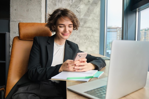 Imagen de una joven vendedora con traje trabajando en una computadora portátil sentada en su oficina con un teléfono inteligente respondiendo