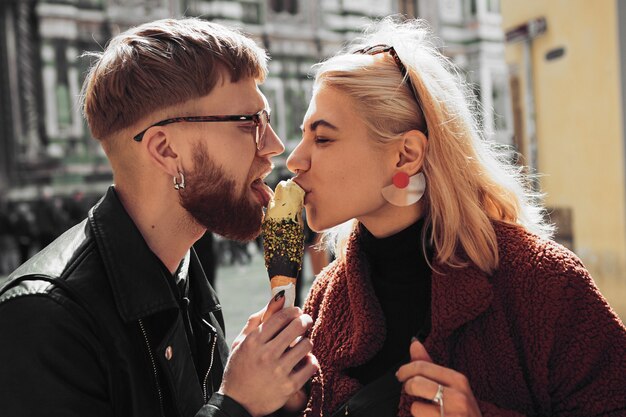 Imagen lateral de una mujer joven rubia con un elegante pendiente y un abrigo de felpa y un hombre barbudo de moda con una chaqueta de motorista y gafas comiendo juntos un helado en la calle