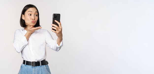 Imagen de una linda chica asiática chateando en video enviando un beso de aire a la cámara tomando selfie con filtros de aplicaciones en el teléfono inteligente de pie sobre fondo blanco