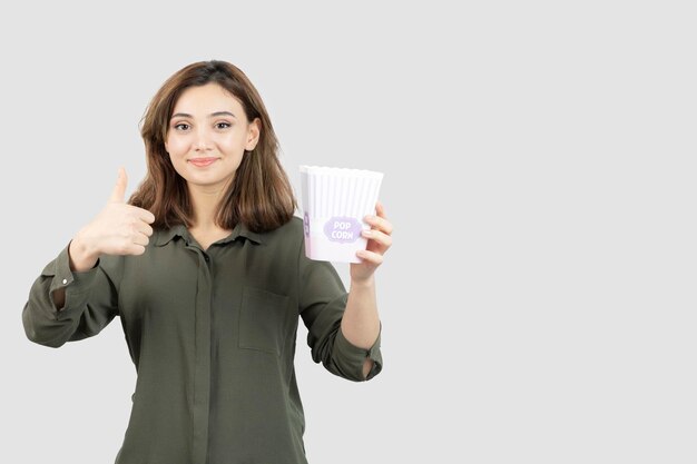 Imagen de linda chica con caja de palomitas de maíz posando para la cámara y dando pulgar hacia arriba. Foto de alta calidad