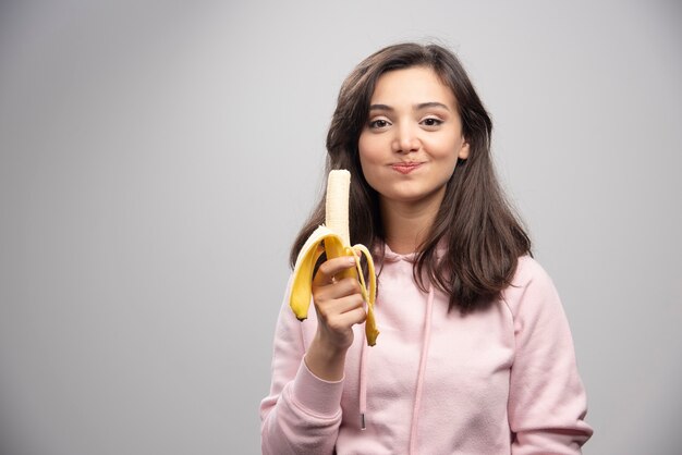 Imagen de una linda chica modelo comiendo un plátano en la pared gris