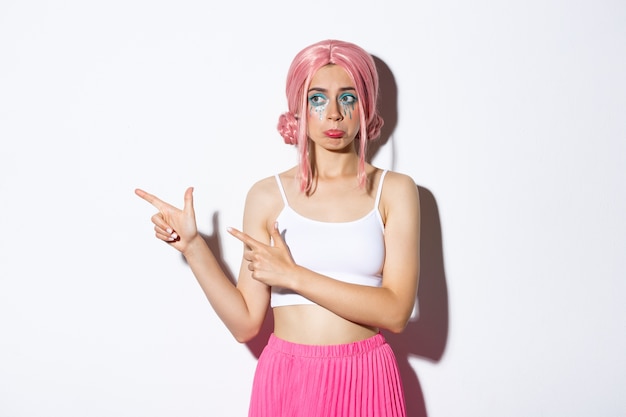 Imagen de una linda niña triste con peluca rosa y maquillaje de halloween, enfurruñada molesta y frunciendo el ceño, señalando con el dedo a la izquierda algo decepcionante, de pie.