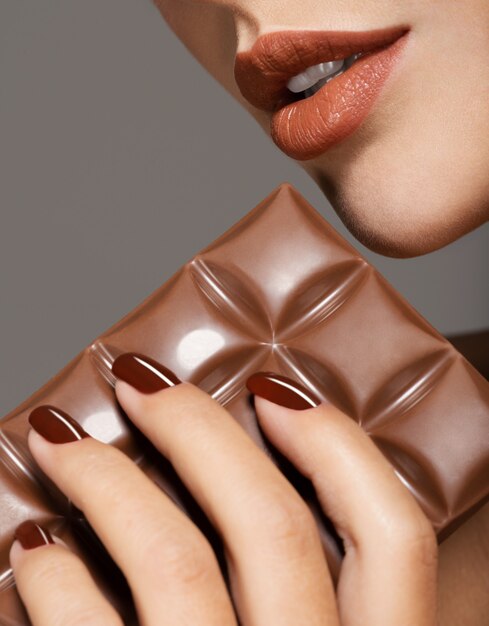 Imagen macro de mano femenina con uñas marrones y barra de chocolate