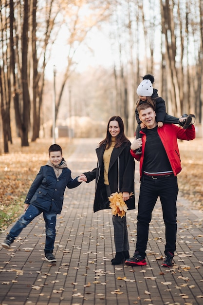 Imagen de mamá con cabello largo y negro con abrigo negro, papá con cabello corto en chaqueta roja, niño bonito con su hermana menor sosteniendo ramos de hojas de otoño