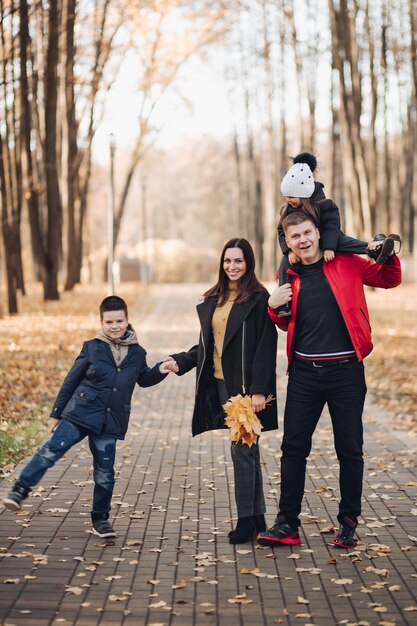 Imagen de mamá con cabello largo y negro con abrigo negro, papá con cabello corto en chaqueta roja, niño bonito con su hermana menor sosteniendo ramos de hojas de otoño