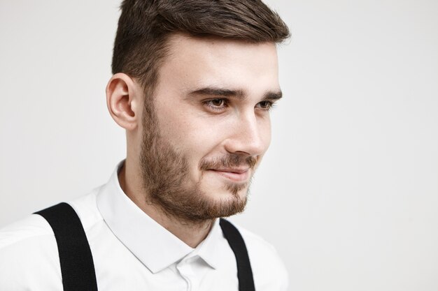 Imagen de medio perfil de un hombre joven de moda de aspecto amistoso con bigote y barba sonriendo pensativamente mientras recuerda alguna historia divertida o broma, posando en el estudio con camisa blanca formal