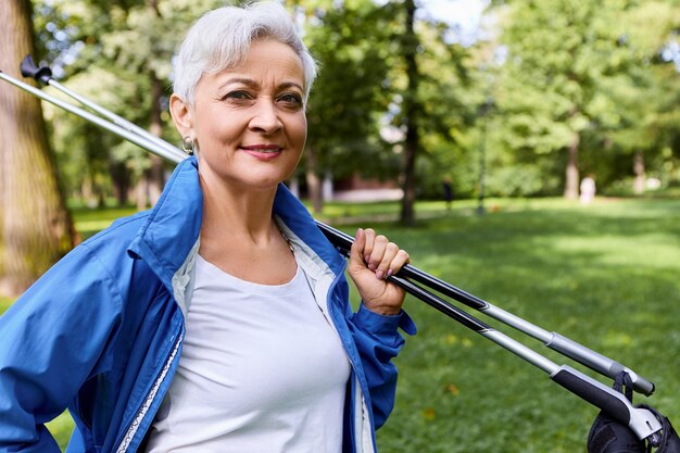 Imagen de moda dama europea confiada con pelo corto gris de pie en un bosque de pinos con bastones de marcha nórdica en los hombros, yendo a casa después del entrenamiento cardiovascular, sonriendo ampliamente