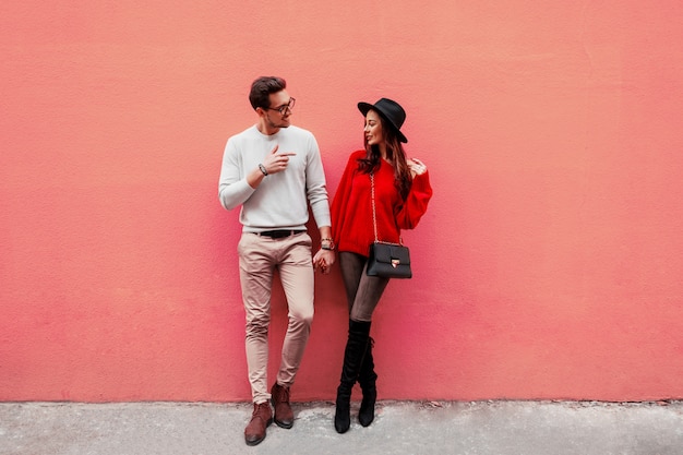 Imagen de moda de elegante elegante pareja de enamorados cogidos de la mano y mirándose con placer. Mujer de pelo largo en suéter de punto rojo con su novio posando.