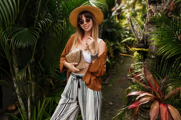 Imagen de moda de mujer elegante sexy con sombrero de paja posando en hojas de palmeras tropicales