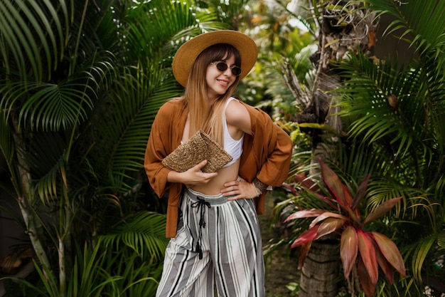 Imagen de moda de mujer elegante sexy con sombrero de paja posando en hojas de palmeras tropicales